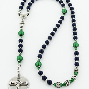 May include: A navy blue and green rosary with a silver crucifix and a silver charm in the shape of a chalice. The rosary beads are made of glass and have a pearl-like finish. The beads are interspersed with green beads that have a metallic finish. The rosary is personalized with the name "CHAD" in silver letters.