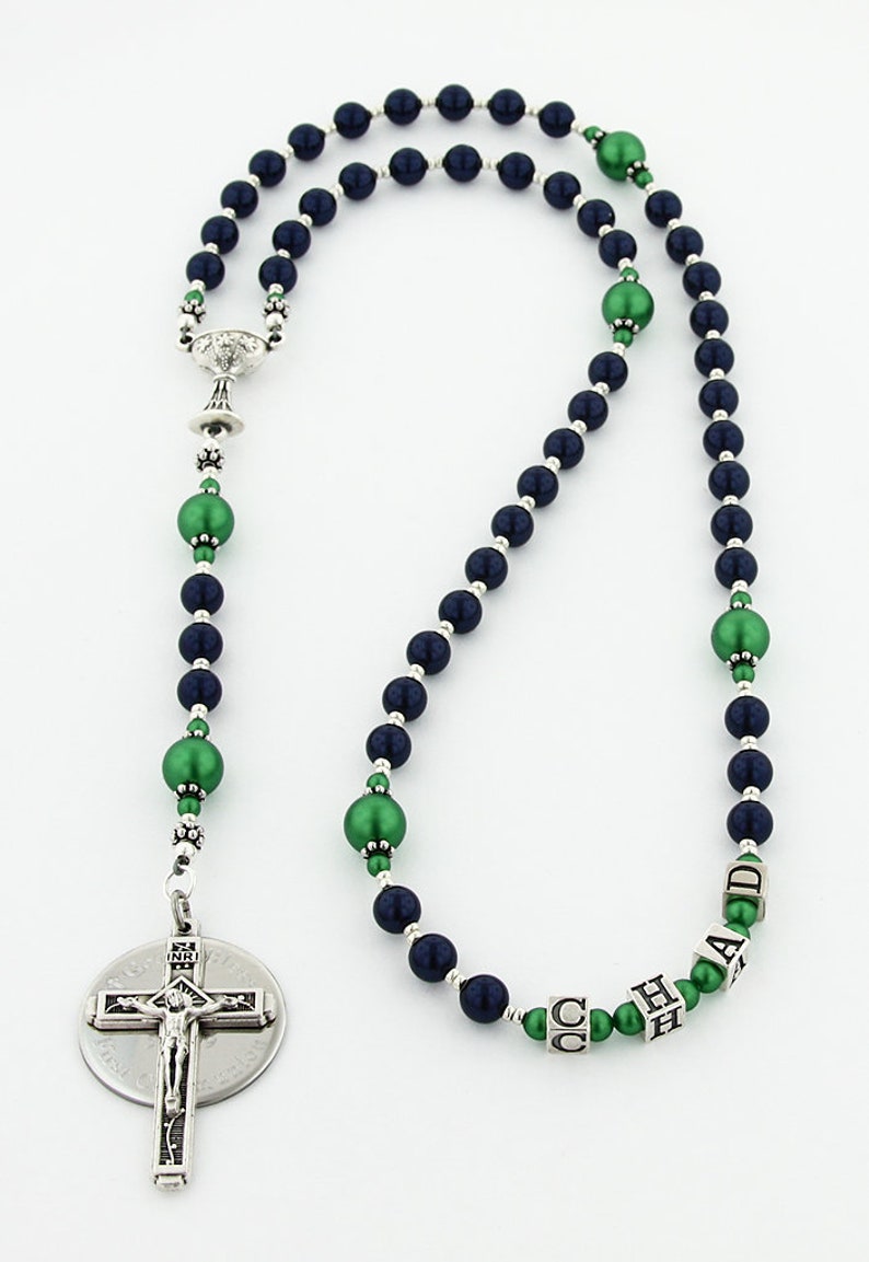 May include: A navy blue and green rosary with a silver crucifix and a silver charm in the shape of a chalice. The rosary beads are made of glass and have a pearl-like finish. The beads are interspersed with green beads that have a metallic finish. The rosary is personalized with the name "CHAD" in silver letters.