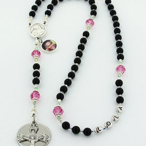 May include: Black rosary beads with a silver crucifix and a silver charm with a photo of a young girl. The beads are interspersed with pink and green crystal beads. The charm also has a silver medal of the Virgin Mary. The rosary is personalized with the name "LOLA" on the beads.