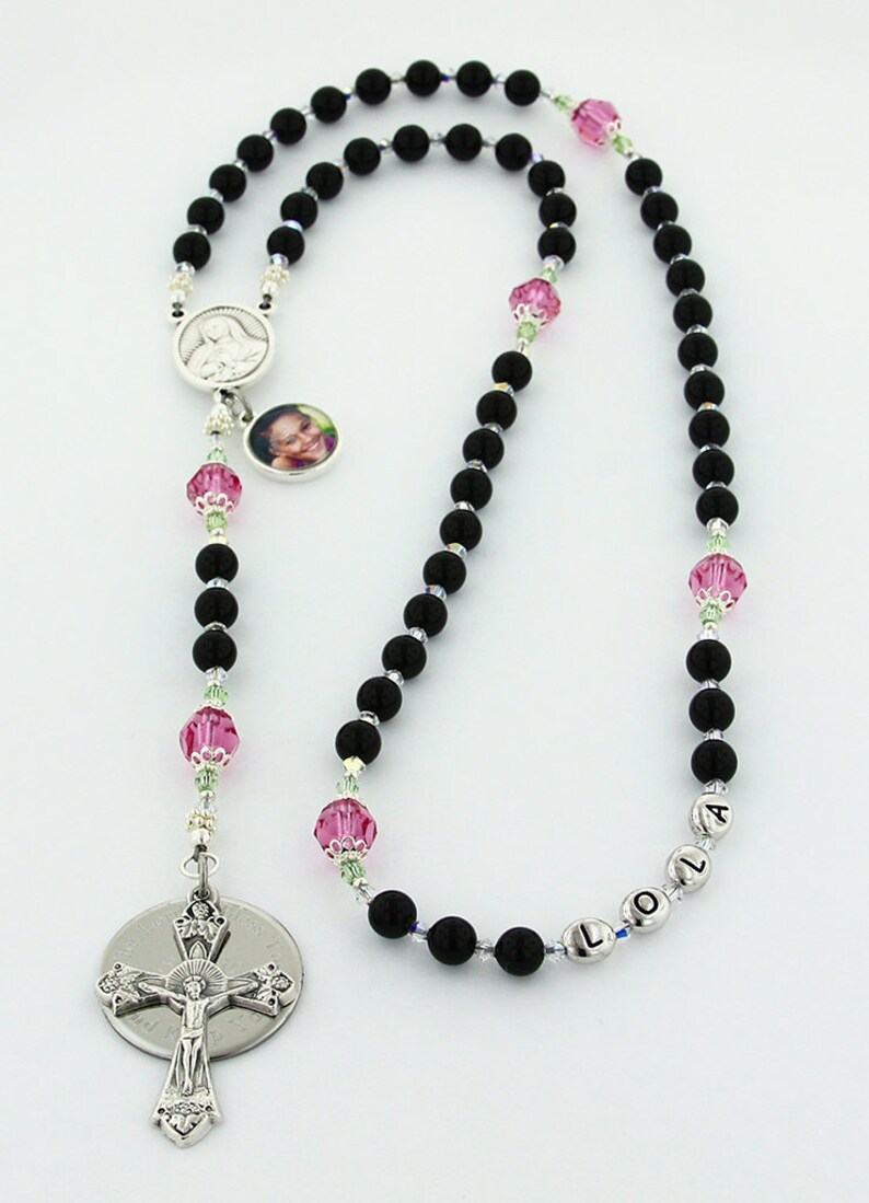 May include: Black rosary beads with a silver crucifix and a silver charm with a photo of a young girl. The beads are interspersed with pink and green crystal beads. The charm also has a silver medal of the Virgin Mary. The rosary is personalized with the name "LOLA" on the beads.