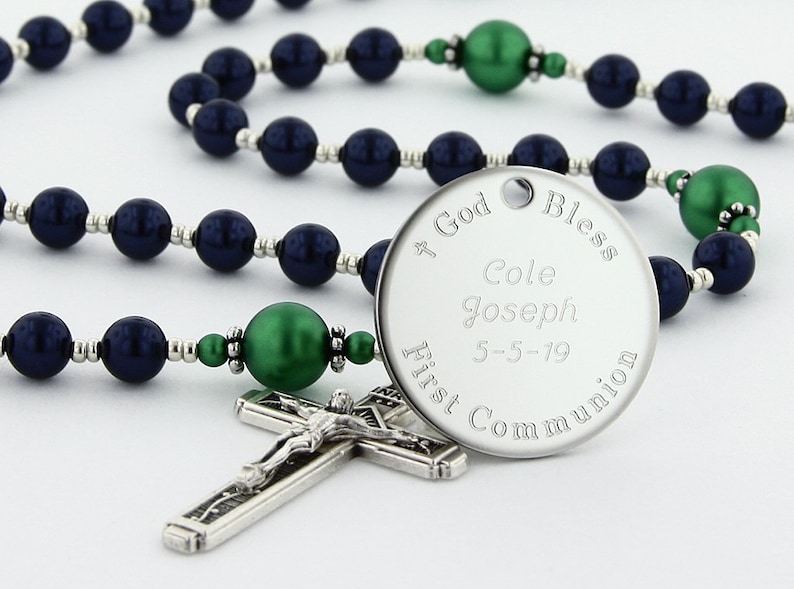 May include: A silver rosary with dark blue and green beads. The center piece is a round silver charm with the inscription "God Bless Cole Joseph 5-5-19 First Communion".