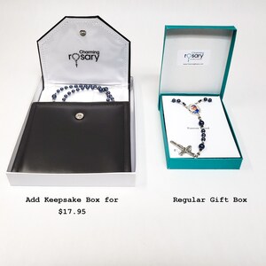 May include: Two gift boxes for a rosary. The box on the left is a keepsake box with a black velvet interior. The box on the right is a regular gift box with a teal blue exterior. Both boxes have the text "Charming Rosary" on them.