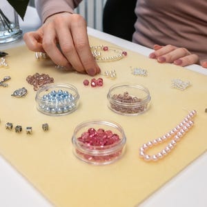 May include: A close-up of a craft table with various beads, containers, and tools for making jewelry. There are pink, blue, and white beads, as well as silver charms and pliers.