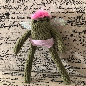 May include: A green knitted fairy doll with pink wings and a pink tutu. The doll has a pink flower on its head and a friendly expression.