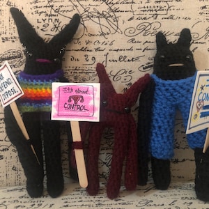 May include: Four crocheted figures holding signs with different messages. The signs read "Deny, Defend, Dispose", "It's about control", "I am confused", and a sign with a uterus symbol. The figures are black, maroon, and blue.