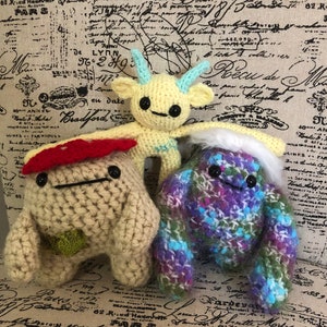 May include: Three handmade crochet creatures. One is yellow with blue horns, another is beige with a red mushroom cap, and the third is a mix of purple, blue, and green with white fur. They have black button eyes and are on a patterned fabric.