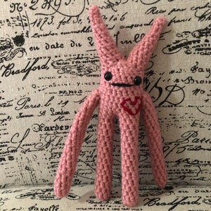 Op de afbeelding: Een roze gehaakt amigurumi-speelgoed met lange ledematen en een rood hart op de borst. Het heeft zwarte ogen en een zwarte mond.
