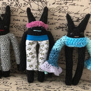 May include: Three black crocheted bunny plushes with different outfits. The first bunny is wearing a silver sequined dress. The second bunny is wearing a blue headband, a blue belt, and white pants with gold stars. The third bunny is wearing a light blue and green crocheted scarf.
