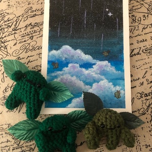 May include: Three green crocheted plushies with leaf-shaped wings. The plushies have black eyes and a smiling mouth. The plushies are in front of a painting of a night sky with clouds and stars.