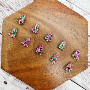 May include: A collection of small, glittery Christmas tree-shaped ornaments in shades of pink, green, and gold. The ornaments are arranged on a wooden, hexagonal surface with a natural wood grain finish. The background is a light-colored wooden surface.