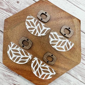 May include: Four pairs of half-circle earrings with a white leaf pattern on a clear background. Each earring has a brown wooden post.