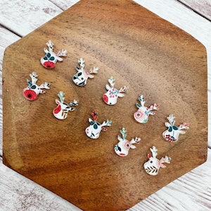 May include: A collection of small, decorative reindeer-shaped embellishments. Each reindeer has a floral pattern in various colors, including red, white, and green. The embellishments are arranged on a wooden surface.