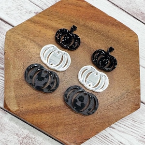 May include: A collection of pumpkin-shaped charms in various styles. Some are black with glitter, others are clear, and some feature a leopard print design. The charms are arranged on a wooden surface.