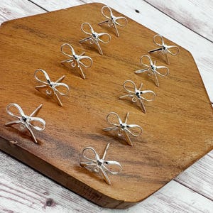 May include: A collection of silver-colored jewelry findings, each with a bow-like design and multiple prongs, are arranged on a wooden, hexagonal surface. The findings are displayed against a backdrop of light-colored wood.