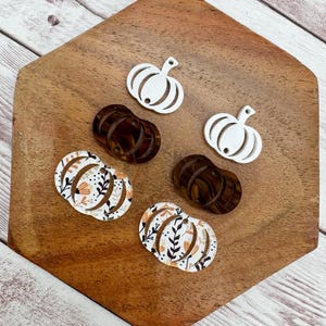 May include: A collection of fall-themed pumpkin cutouts in various designs. There are two white pumpkins, two brown pumpkins, and two pumpkins with a floral pattern in orange, brown, and white. The cutouts are arranged on a wooden surface.