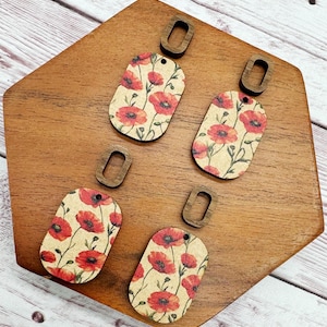 May include: Five wooden poppy flower earrings with matching wooden earring hooks. The earrings have a beige background with red poppy flowers and green leaves. The earring hooks are a dark brown color. The earrings are on a wooden surface.