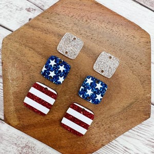 May include: Four square-shaped glitter embellishments with different designs. Two are red and white stripes, one is blue with white stars, and one is white with silver glitter.