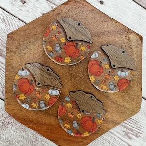 May include: Four unfinished wooden pumpkin-shaped ornaments with a fall-themed design. The ornaments are painted with orange, white, and yellow pumpkins, leaves, and acorns. The ornaments are on a wooden surface.
