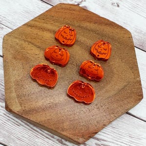 May include: Six orange, reflective pumpkin-shaped charms with carved faces are arranged on a wooden, hexagonal tray. The pumpkins are a bright, glossy orange, perfect for Halloween-themed crafts or decorations.
