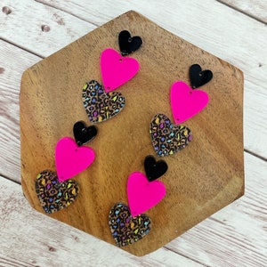 May include: Set of six layered heart-shaped acrylic charms. Each charm features a pink heart layered on top of a leopard print heart. The top layer of each charm is a black heart.
