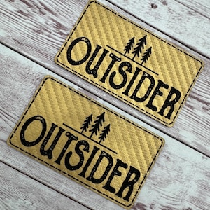May include: Two rectangular patches with a gold woven texture. The word "OUTSIDER" is printed in black, with a line and three pine trees above the word. The patches have a black stitched border.
