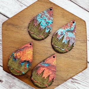 May include: Four teardrop-shaped earrings with a nature-inspired design. Each earring features a landscape scene with a forest silhouette and a colorful sky in shades of orange, blue, and red. The earrings are set on a wooden surface.