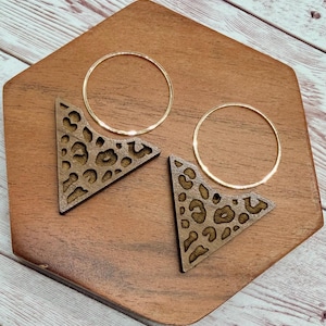 May include: A pair of earrings featuring thin gold-tone circular hoops attached to triangular wooden pendants. Each wooden piece displays a laser-cut leopard print pattern with dark brown spots against a lighter brown wood grain background.