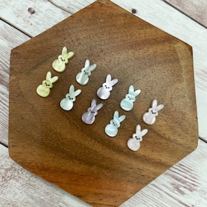 May include: A collection of pastel-colored bunny shaped buttons with a pearlescent finish. The buttons are arranged on a wooden surface.