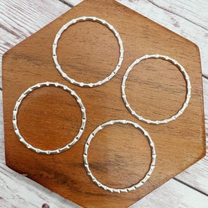 May include: Four silver-colored, textured metal rings with a repeating geometric pattern. The rings are arranged on a dark brown, hexagonal wooden surface. The rings are circular and appear to be jewelry components or decorative elements.