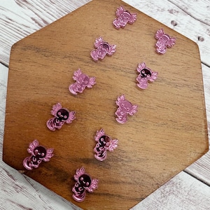 May include: Pink axolotl-shaped acrylic charms scattered on a wooden surface. Each charm features a detailed axolotl design with wing-like appendages. The charms are reflective and appear to be made of a shiny material.