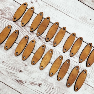 May include: Twenty wooden oval-shaped tags with two holes each. The tags are a light brown color and are arranged in two rows on a white wooden surface.
