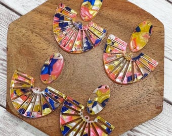 May include: A set of six colorful acrylic earrings. The earrings are in the shape of a fan and an oval. The fan earrings have a gold and white pattern. The oval earrings have a pink, blue, and yellow pattern.