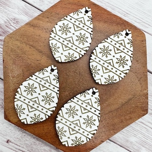May include: Four white teardrop-shaped wooden earring blanks with a laser-etched geometric pattern featuring snowflakes and zigzags. Each blank has a small hole at the top for attaching earring hooks. The earrings are displayed on a wooden tray.