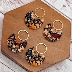 May include: Four crescent-shaped charms featuring a dark floral pattern with yellow, white, and muted blue flowers on a black background. Each charm is attached to a thin, circular gold-toned metal ring, creating a set of decorative accessories.