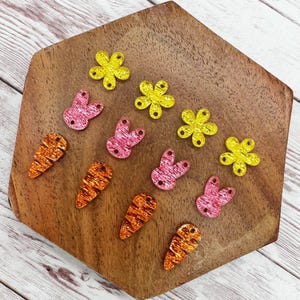 May include: Assortment of glitter Easter-themed craft supplies.  There are four pink bunny shapes, four orange carrot shapes, and four yellow flower shapes.