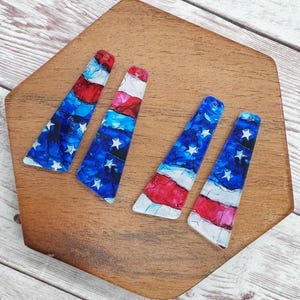 May include: Four patriotic-themed earrings with a unique design, featuring a blend of red, white, and blue colors. The earrings are shaped like elongated triangles, with a pattern of stars and stripes. They are displayed on a wooden surface.
