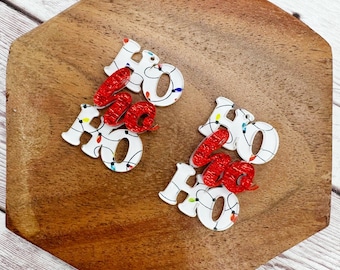 May include: Two sets of Christmas-themed earrings spelling "HO". The white letters are decorated with colorful lights, and the "HO" is accented with red glitter. The earrings are displayed on a wooden surface.