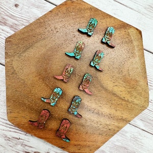 May include: A collection of nine wooden cowboy boot charms with a turquoise and rust colored paint design. The charms are arranged in a pattern on a wooden surface.