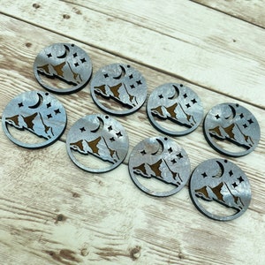 May include: Eight wooden round ornaments with a crescent moon and stars design. The ornaments are painted in a silver color with a brown mountain silhouette.