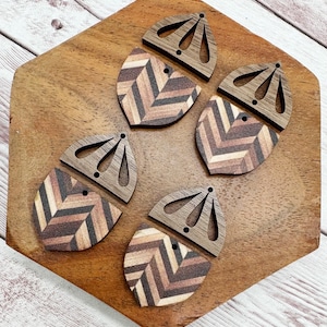 May include: Six wooden earring components with a geometric chevron pattern in shades of brown, beige, and black. The top portion features a decorative cutout design. The earrings are displayed on a wooden surface.