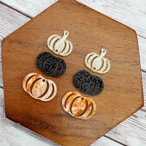 May include: A collection of pumpkin-shaped charms in various colors and textures. There are gold glitter, black lace, and orange marbled designs. Each charm has a small hole at the top for stringing. The charms are displayed on a wooden surface.