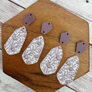 May include: Earring components in the image include four white teardrop-shaped pieces with a floral pattern in mauve and four mauve geometric shapes. The components are arranged on a wooden surface with a hexagonal shape.
