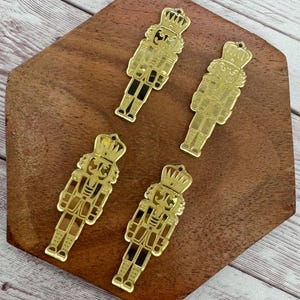 May include: Four gold-colored nutcracker ornaments with a crown and uniform design. The ornaments are arranged on a wooden surface with a dark brown color. The nutcrackers have black accents and small holes for hanging.