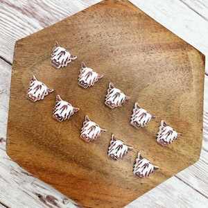 May include: Ten white and brown Highland cow head buttons with glitter accents. The buttons are arranged in a circle on a wooden surface.