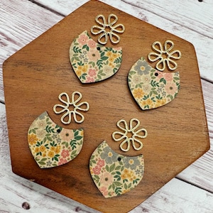 May include: Four wooden earring components with floral patterns in pink, yellow, and green, paired with gold-colored metal flower-shaped connectors. The earrings are arranged on a wooden surface, showcasing a craft or jewelry-making theme.