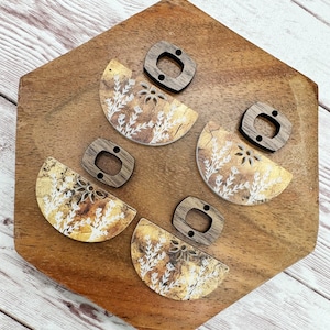 May include: Four pairs of earrings with a gold and white floral pattern on a wooden surface. Each earring is a half circle with a wooden accent piece.