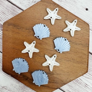 May include: A collection of seashell and starfish charms in white and light blue, arranged on a wooden surface. The charms include five-pointed starfish and fan-shaped seashells, each with a small hole for stringing.