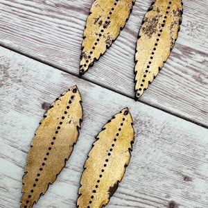 May include: Four gold-colored, leaf-shaped wooden cutouts with a distressed finish. Each cutout has a series of small, evenly spaced black dots running down the center. The edges are dark brown. The cutouts are arranged on a light wood-grain surface.