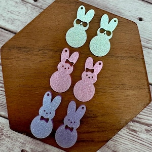 May include: Six glittery bunny-shaped ornaments in shades of green, pink, and purple, each with a bow or sunglasses. The ornaments are arranged on a wooden surface, perfect for Easter or spring crafts.