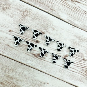 May include: A set of ten cow-print acrylic charms in the shape of a cow's head. The charms are black and white and have a glossy finish.
